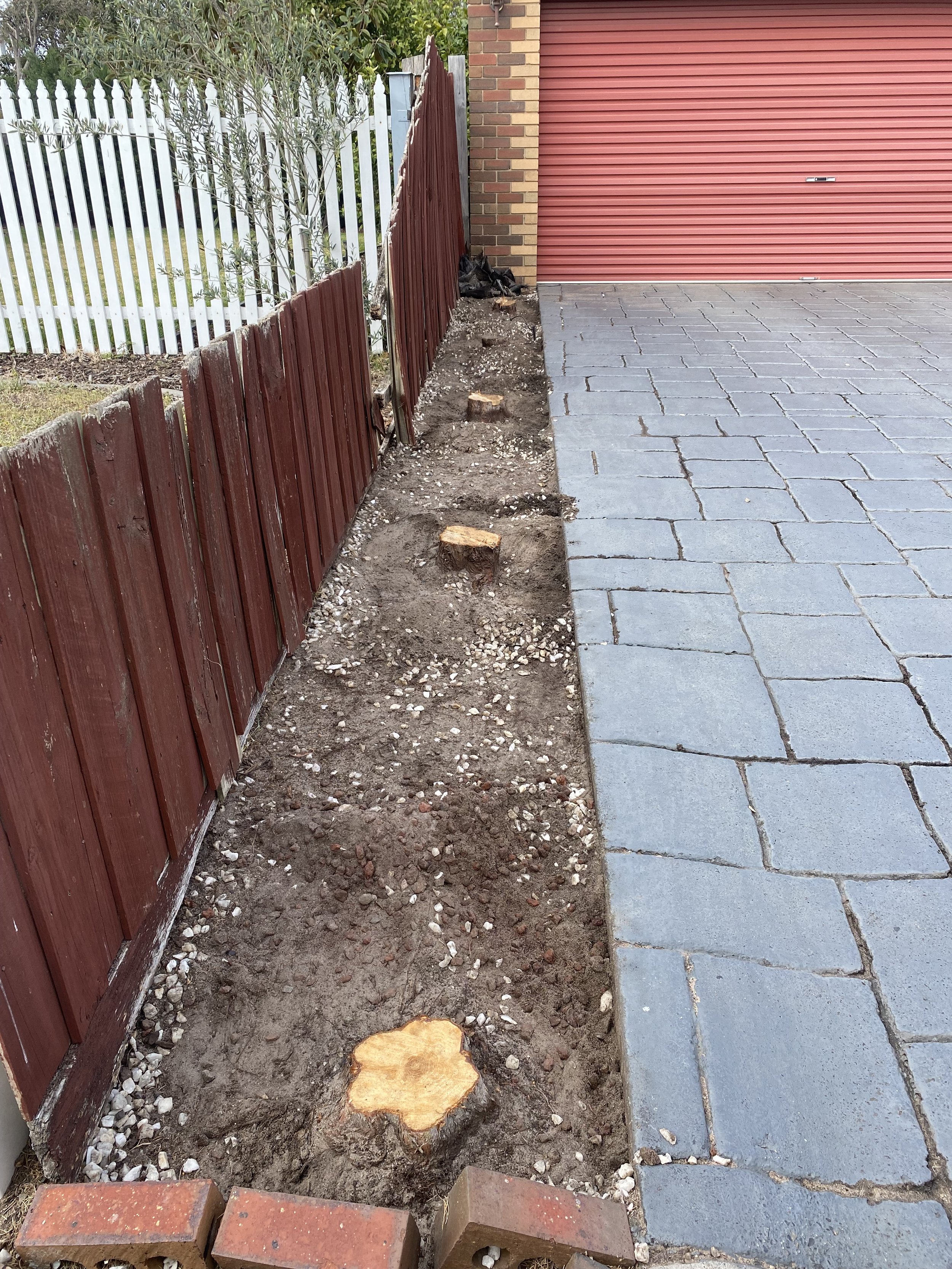 A small garden bed area has been cleared next to a red fence and a brick building with a red roller door. The soil has been loosened, with some wood stumps and a few bricks visible. Part of the stone walkway is on the right side of the image.