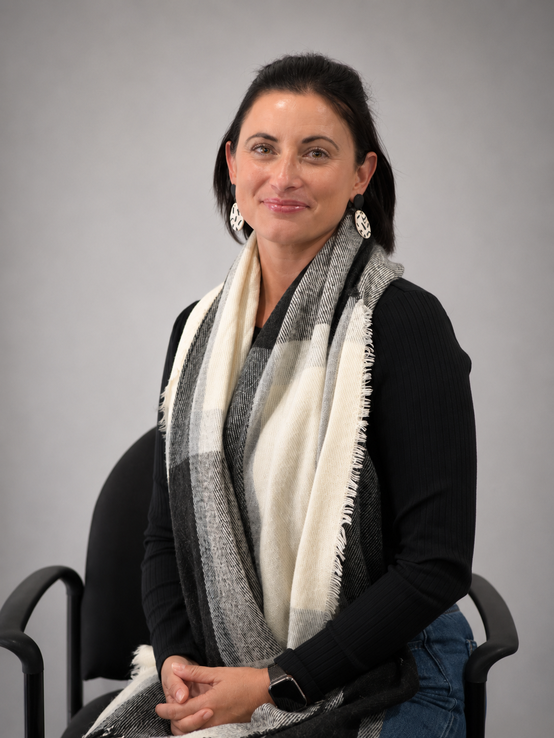 A woman sitting on a black chair with a gray background, wearing a black top, a black and white checkered scarf, and large circular earrings.