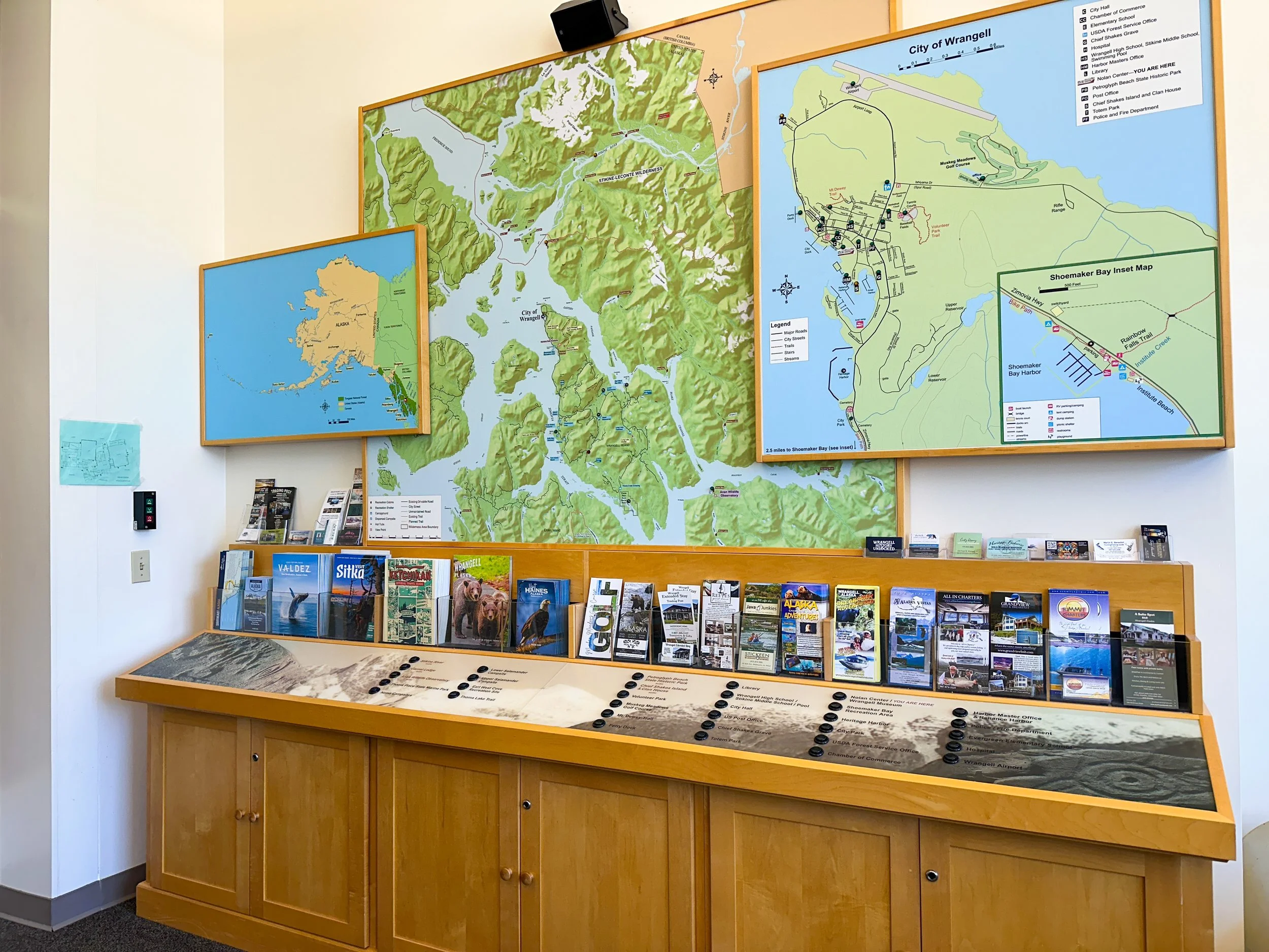 A wall with multiple maps of Wrangell and Alaska, and a brochure display table below with various travel brochures and pamphlets about Wrangell and local attractions.