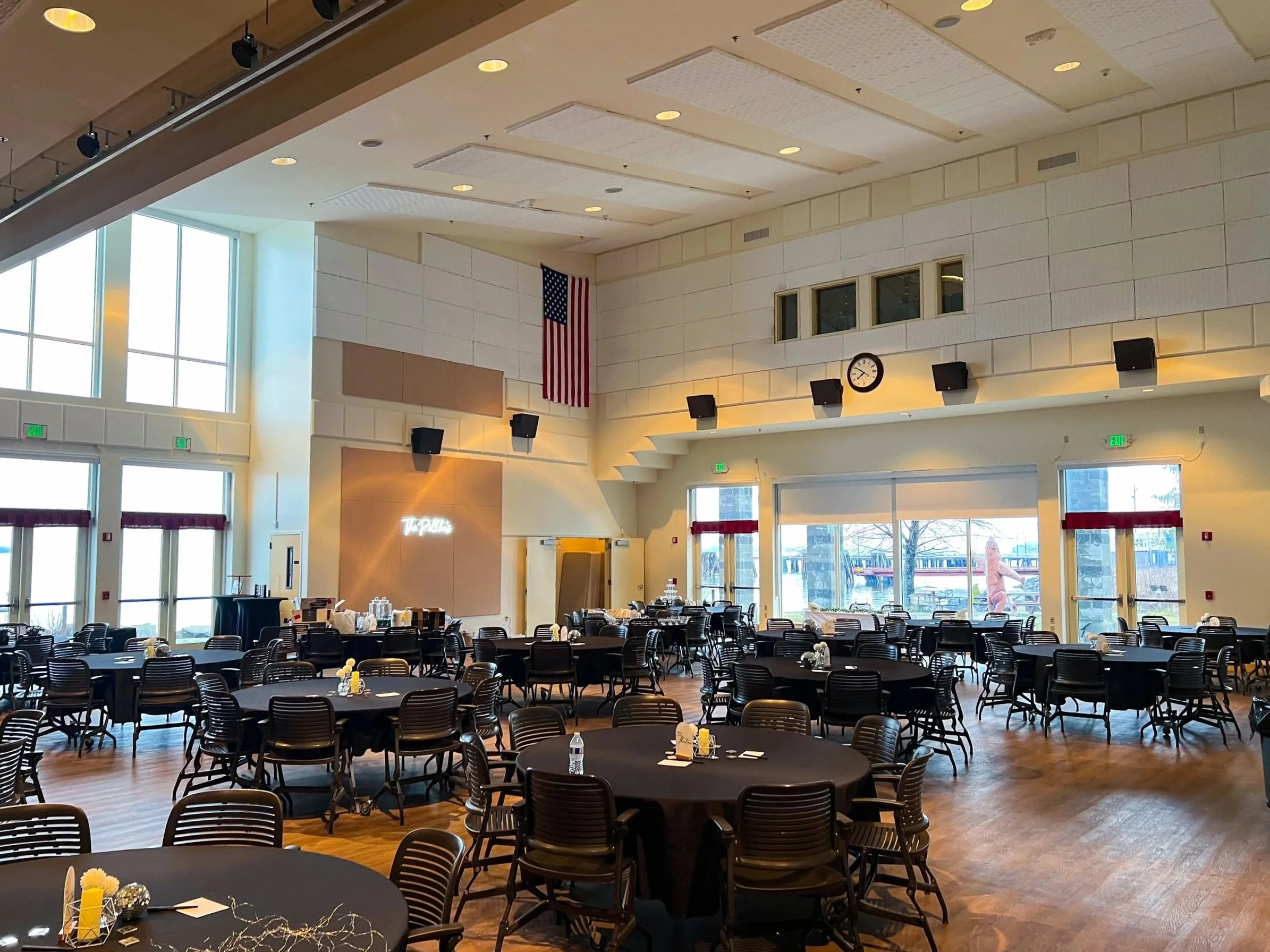 A spacious, well-lit banquet hall set up with round tables covered with black tablecloths, each arranged with place settings and centerpieces. The high ceiling features recessed lighting and acoustic panels. An American flag hangs on the wall, and there is a clock, several speakers, and exit signs visible. Large windows let in natural light, and a person is seen outside near the entrance.