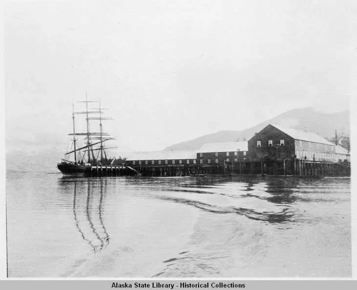 A historical black and white photo of a ship docked at a pier near a large wooden building, with mountains in the background and calm water in the foreground.