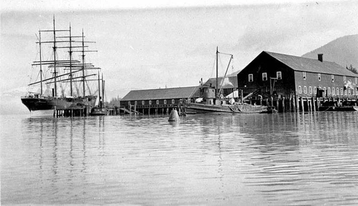 A black-and-white photograph of a harbor with a large sailing ship, a smaller boat, and a long wooden warehouse on stilts over the water, with mountains in the background.