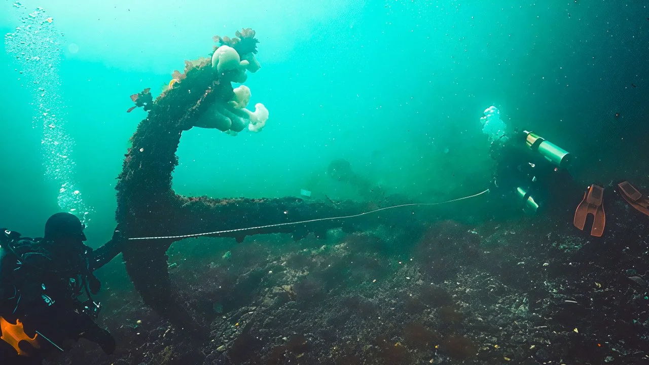 Underwater scene with two scuba divers exploring a sunken shipwreck, one facing the wreck and the other facing away, with bubbles rising and a coral-covered structure.