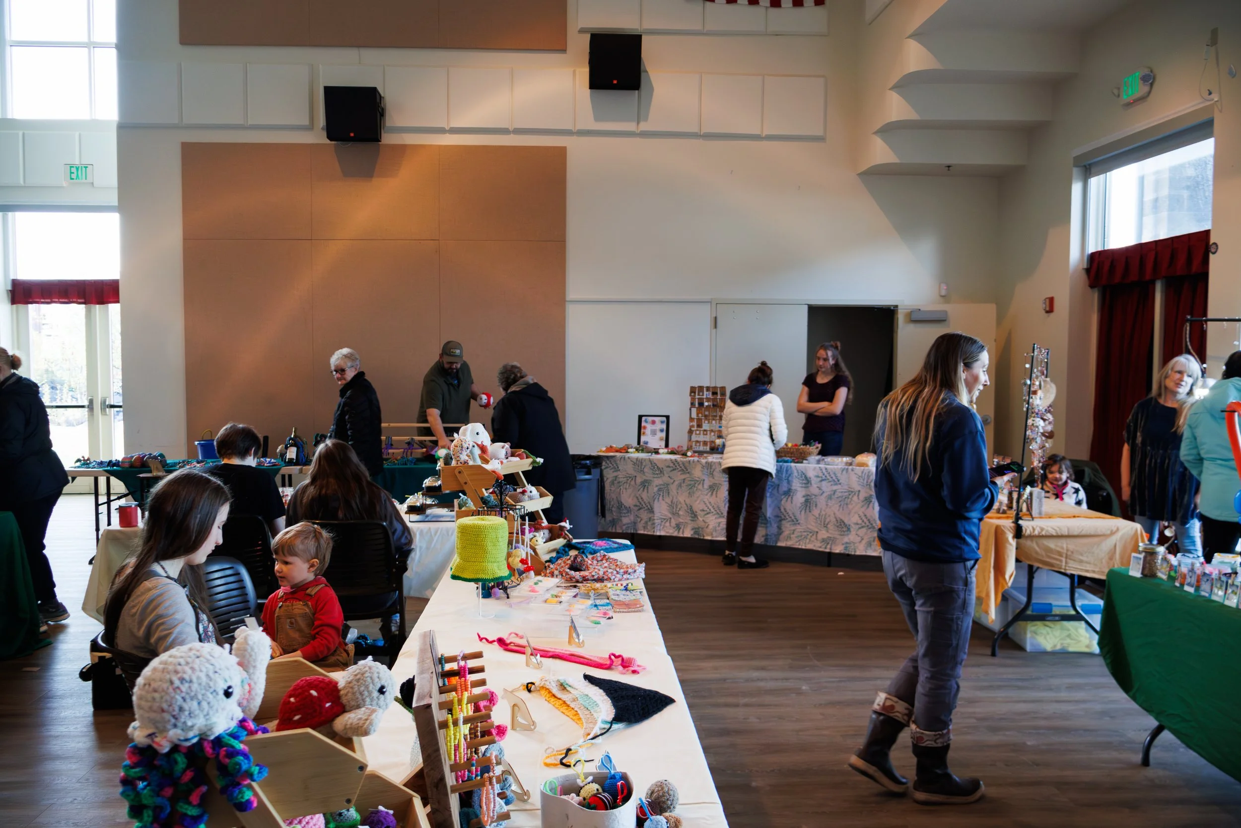 People browsing and selling handmade crafts and knitted items at a craft fair or indoor market, with tables filled with yarn, knitted toys, and accessories in a spacious room with high ceilings and large windows.