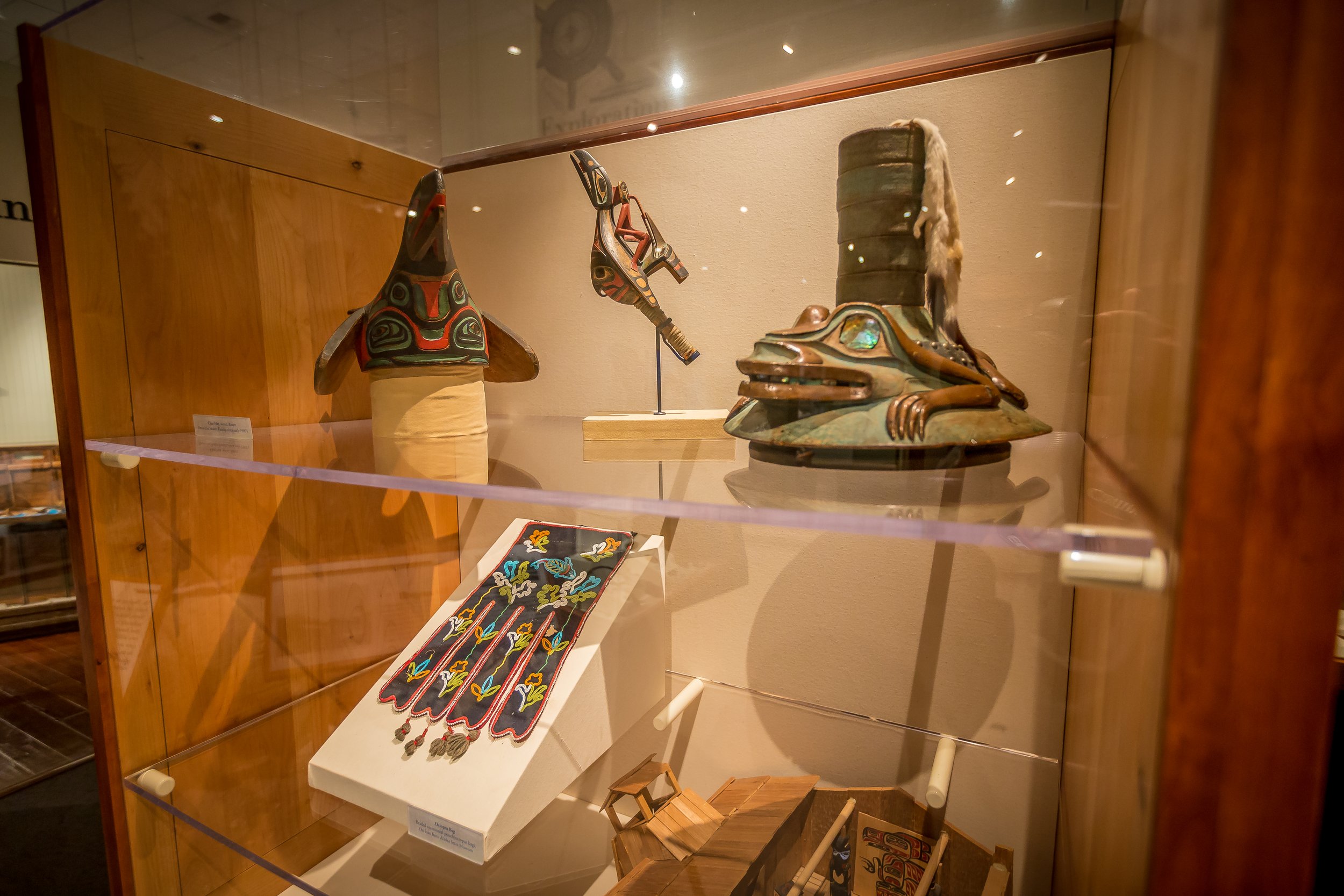 Display case with traditional Native American art and artifacts, including a decorated headdress, a carved wooden figure, embroidered textile, and small wooden items.