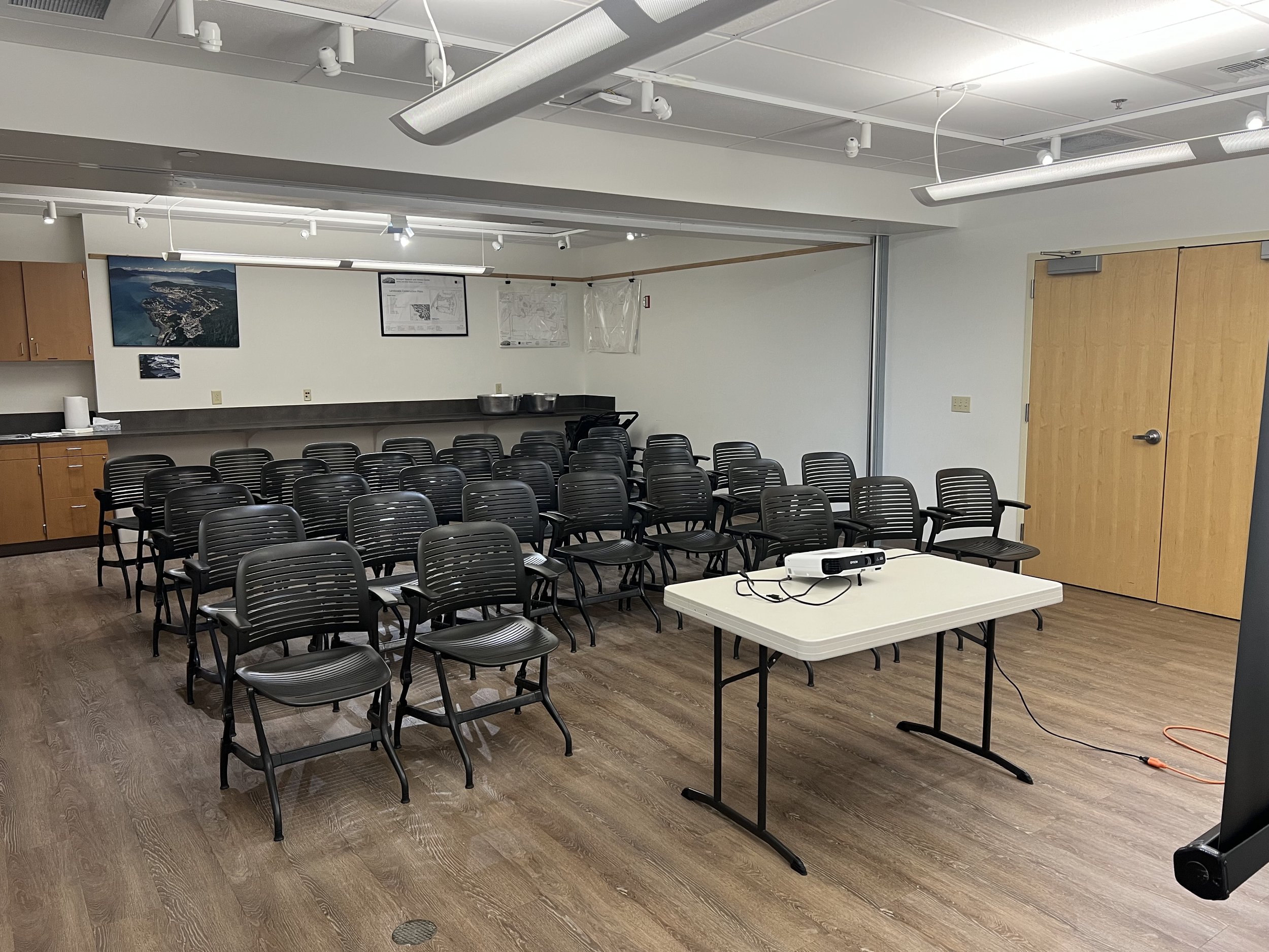 An empty conference or seminar room with many black chairs arranged in rows, a white table in front with a projector on it, and a projection screen mounted on the wall with some posters and pictures nearby. The room has wood flooring, a wooden cabinet on the left, and a closed wooden door on the right.