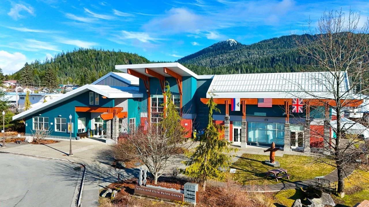 The James and Elsie Notan Center is a modern building with a colorful exterior, featuring red, blue, and gray sections, large glass windows, and flags of the United States, the United Kingdom, and an unspecified country. It is surrounded by trees and landscaped grounds, with mountains and a clear blue sky in the background.