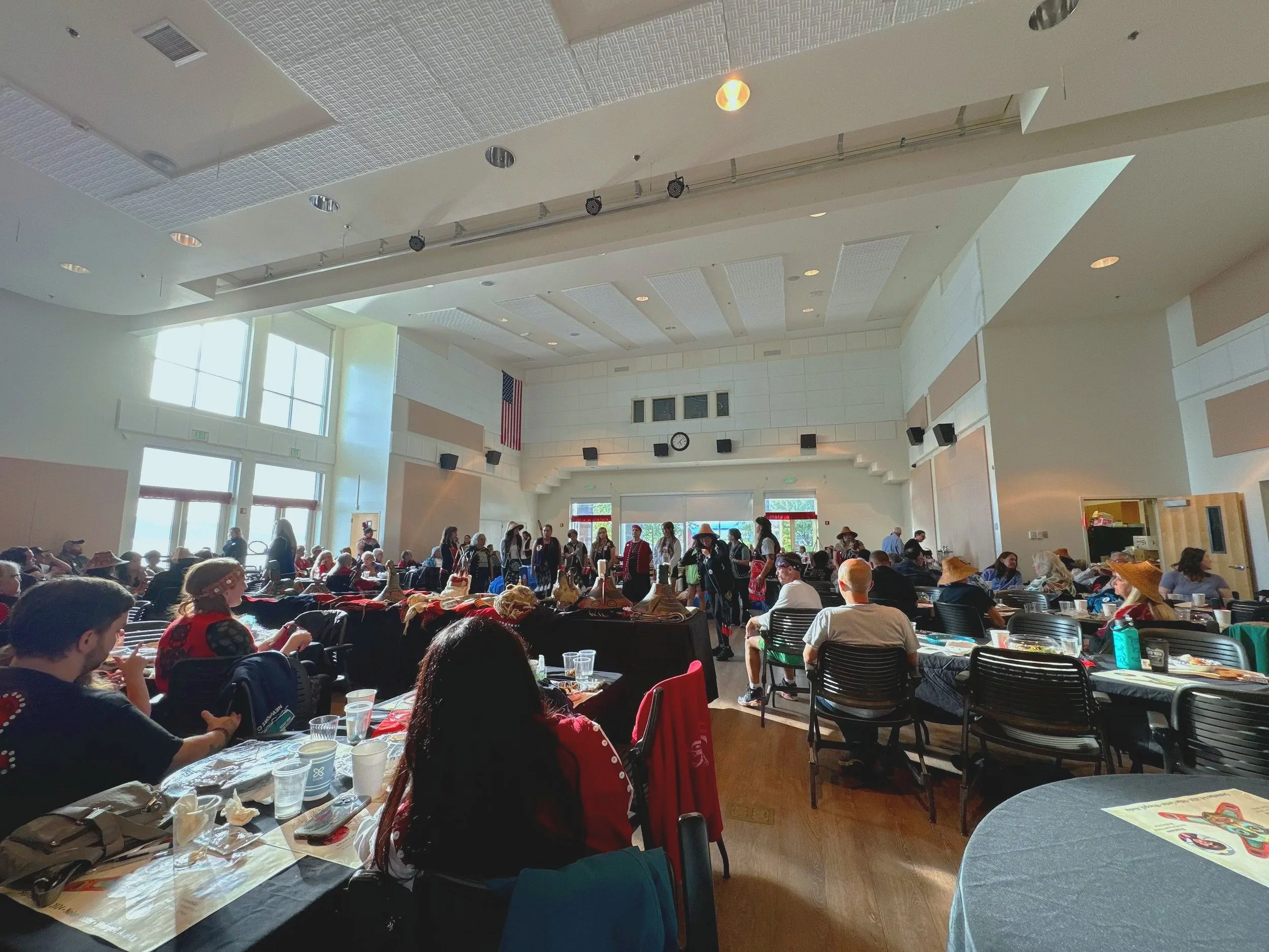 A large indoor gathering with people sitting at tables and some standing, with a high ceiling, large windows, and an American flag on the wall.