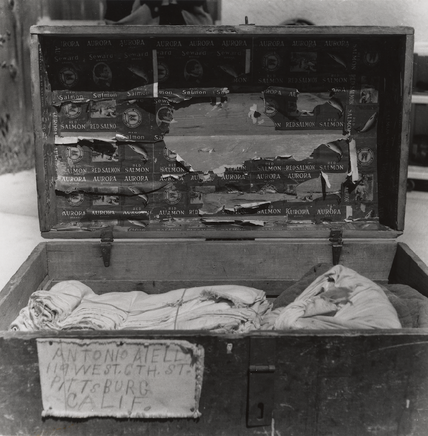 An open wooden box containing rolled cloths or clothing, with a sign labeled 'Antonio Viel, 119 West G. St, Pittsburg, Calif.' The inside of the box's lid is covered in torn and peeling advertisements for salmon.
