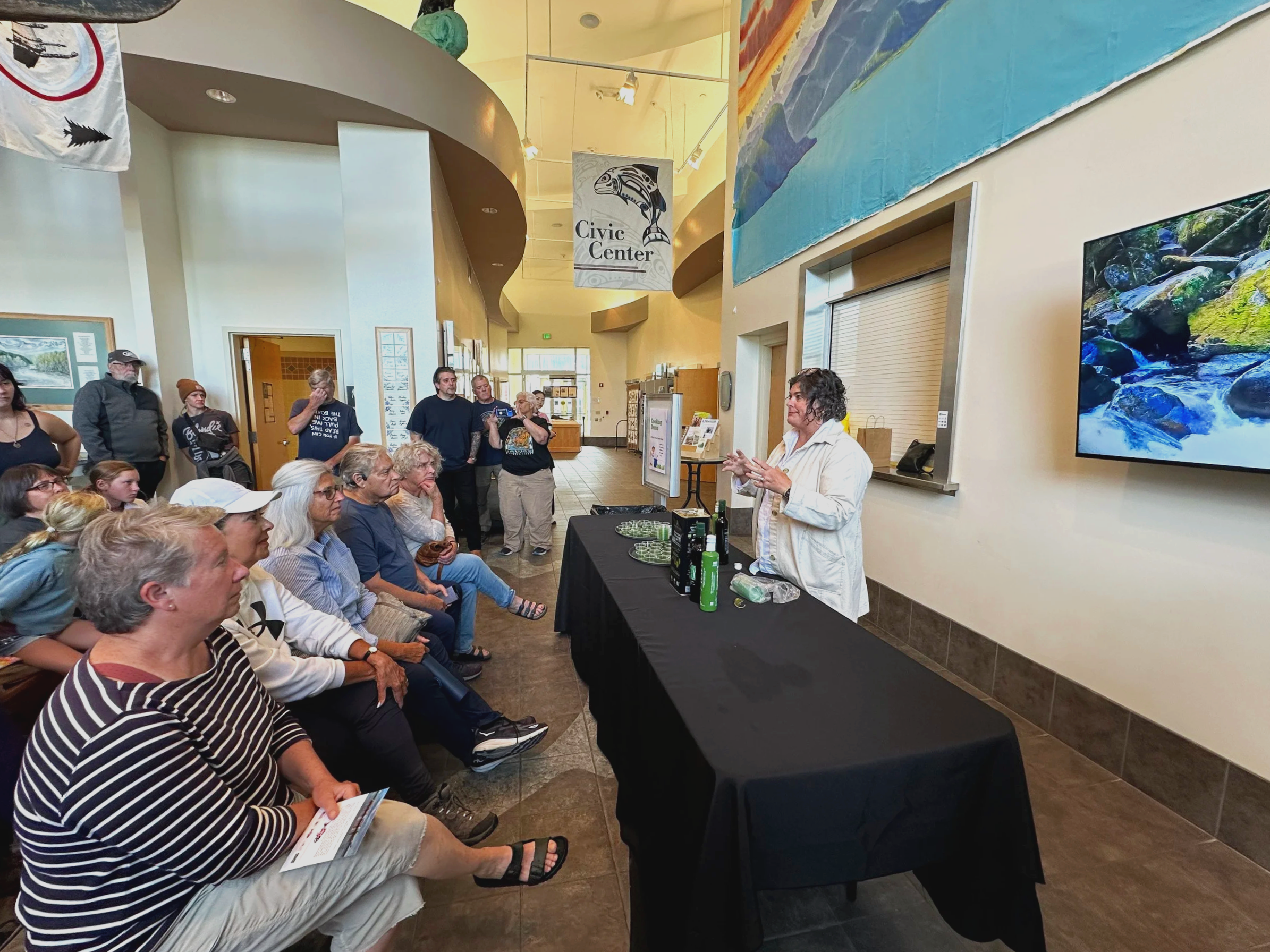 A woman giving a presentation to a seated audience inside a civic center with a large screen displaying a nature scene, and several standing people listening attentively.