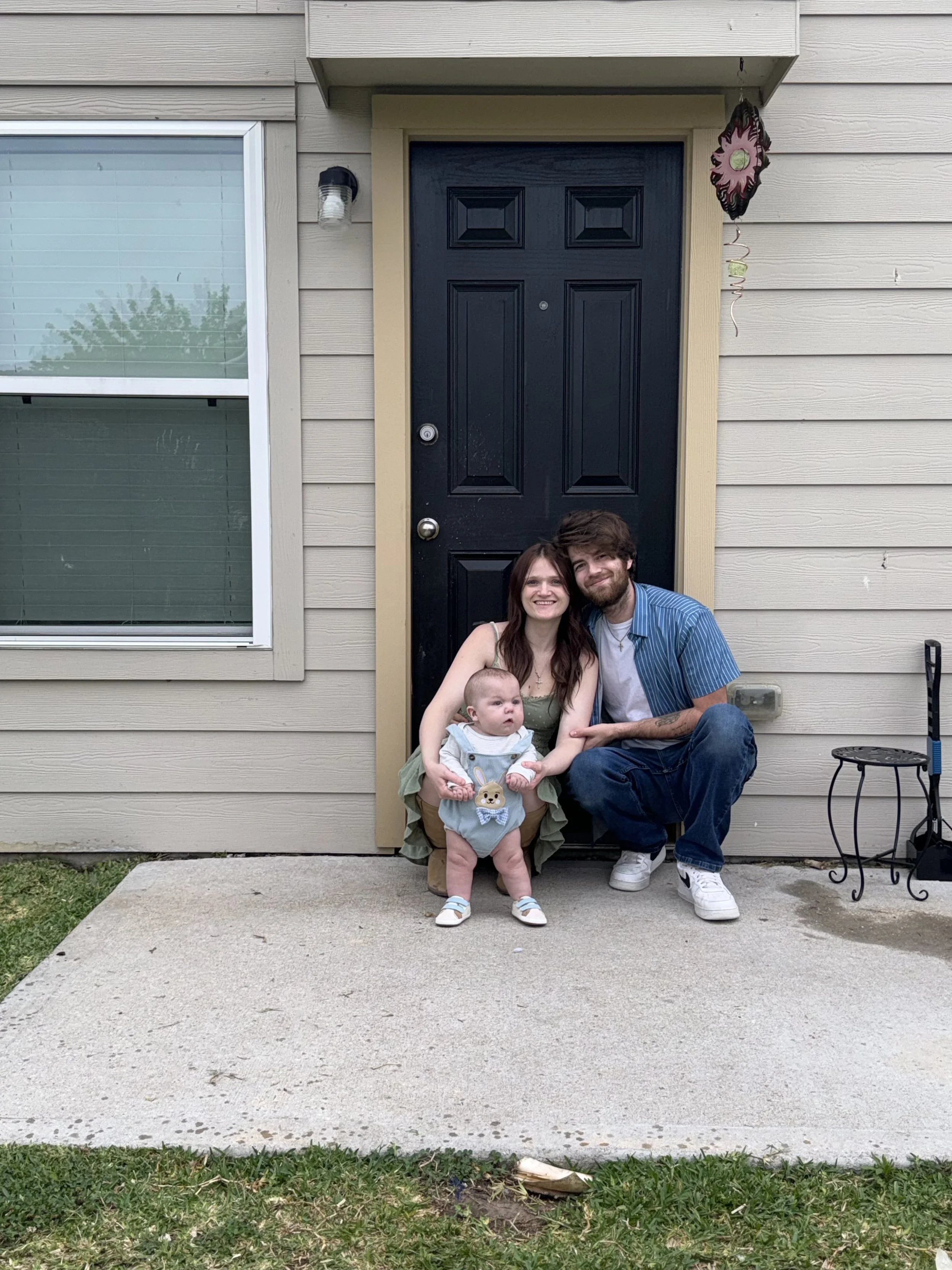 A smiling woman, a smiling man, and a baby girl sitting on the front porch of a house, with the woman holding the baby. The house has beige siding, a black front door, and a window to the left of the door. There is a small black table and a tool on the right side.