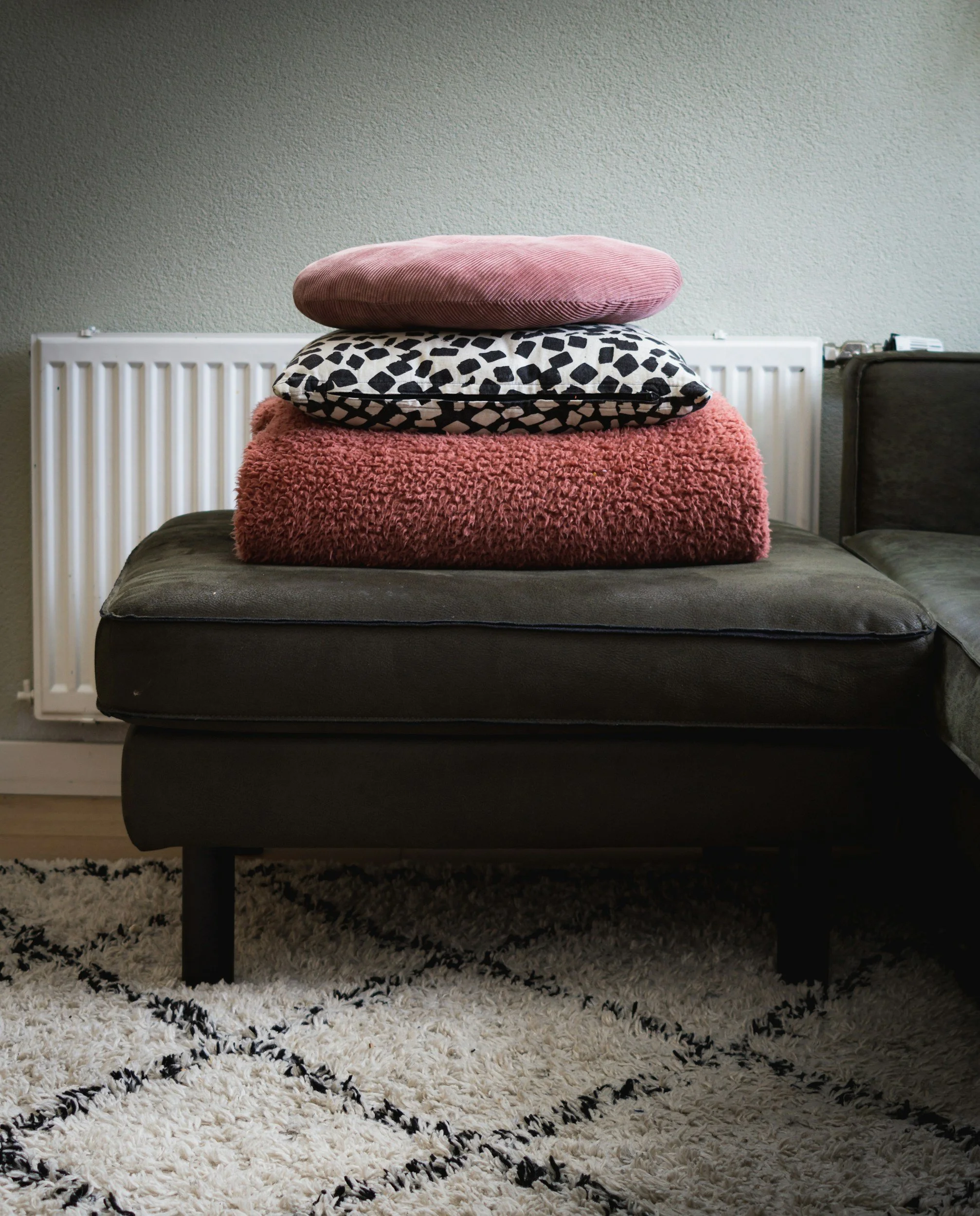 Stack of three cushions on a dark gray sofa, with a textured white and black patterned cushion on top, a pink fluffy cushion in the middle, and a smooth pink cushion on the bottom, in front of a beige wall with a white radiator.