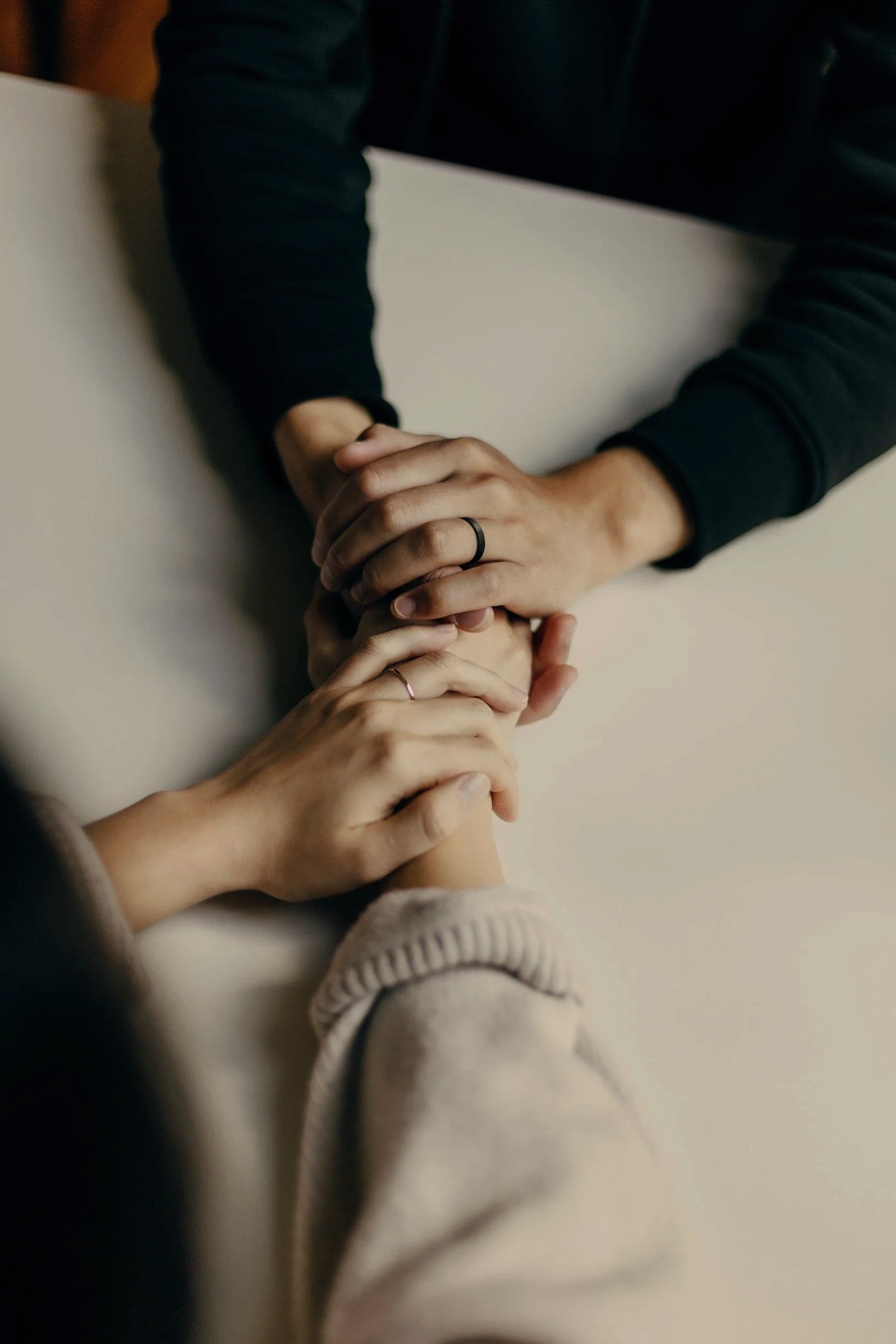 Deux mains qui se tiennent, une personne porte une bague noire et l'autre une bague rose, dans un environnement intérieur.