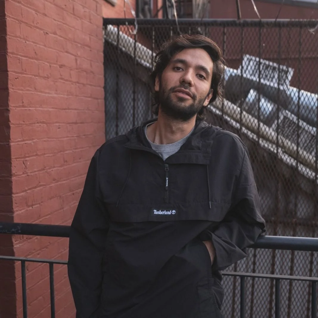 A man with dark wavy hair and a beard leaning against a metal railing on an urban rooftop or fire escape, wearing a black Timberland jacket, with a red brick wall and metal staircase in the background.