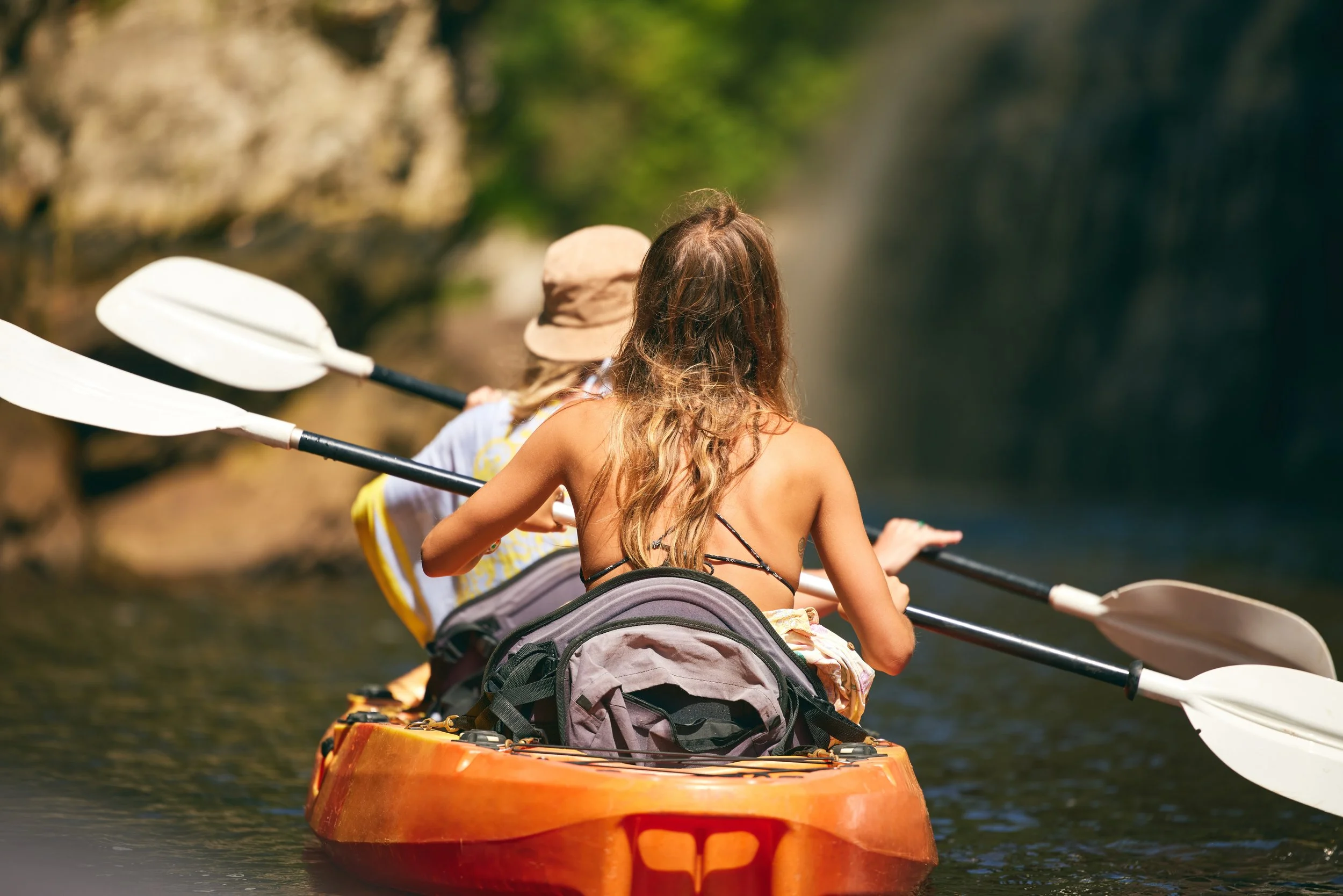 Two women paddling a kayak on a river, surrounded by greenery and rocks.