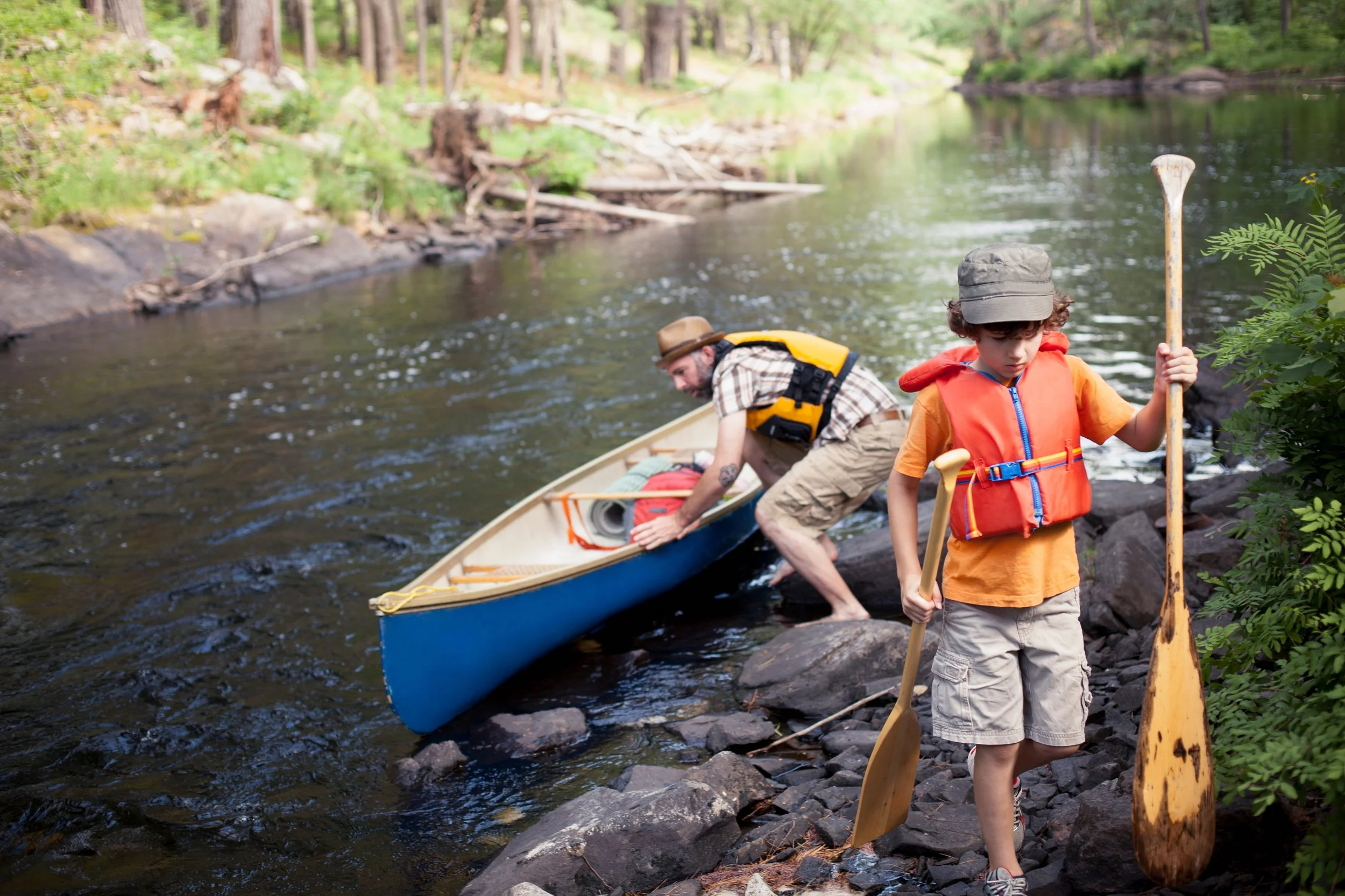 A man and a boy preparing a kayak on the shoreline of a river surrounded by trees. The man is in the water adjusting the kayak, while the boy stands on the rocks holding a paddle and wearing a life jacket.