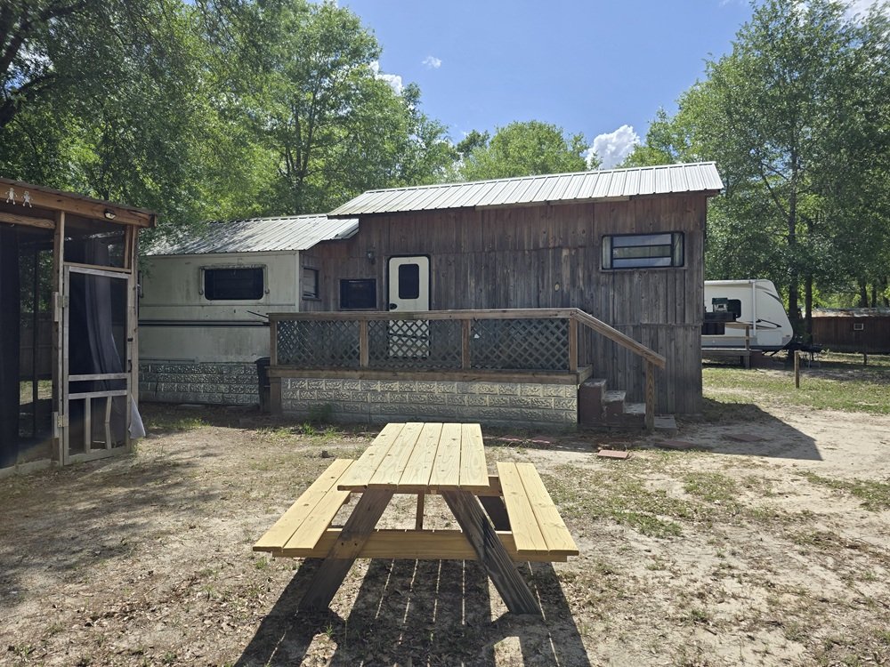 A rustic wooden tiny house with a small porch, dance steps, and a window on a cloudy day, surrounded by trees and a gravel yard with a picnic table in the foreground.