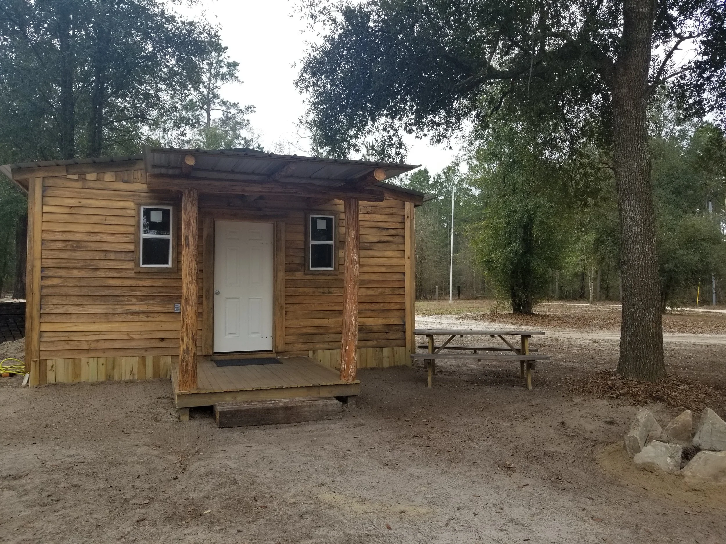 Small wooden cabin with a front porch, two small windows, and a picnic table nearby, surrounded by trees and dirt ground.