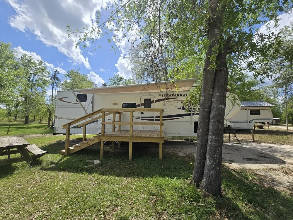 A white travel trailer with wooden steps leading up to the door, set on a grassy area with trees and a picnic table nearby, under a partly cloudy sky.