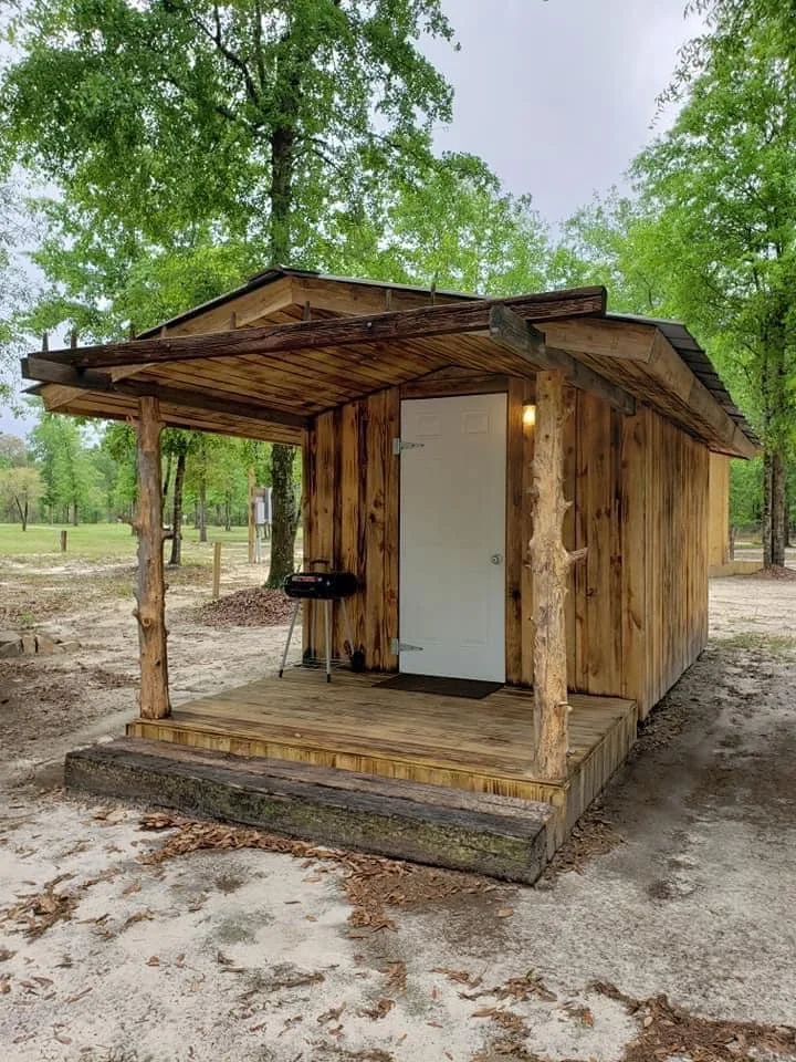 A small wooden cabin with a front porch, white door, and a grill on the porch, surrounded by trees and dirt ground.