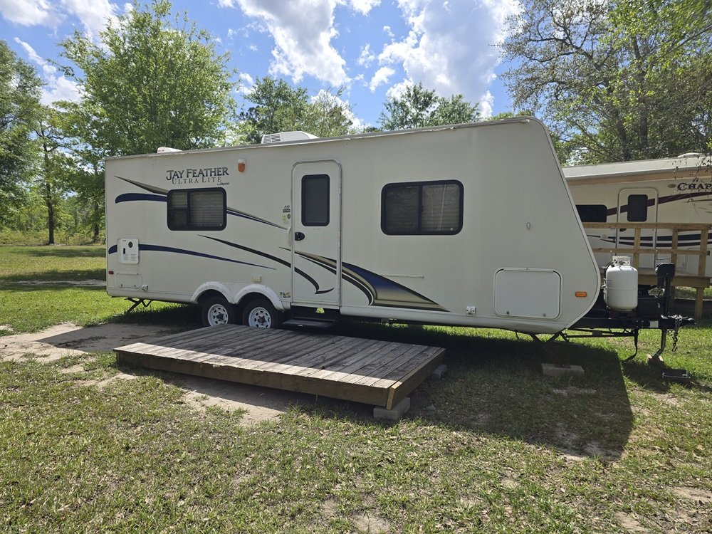A white travel trailer parked on grassy ground with a wooden ramp in front and a second trailer partially visible in the background, surrounded by trees and a partly cloudy sky.