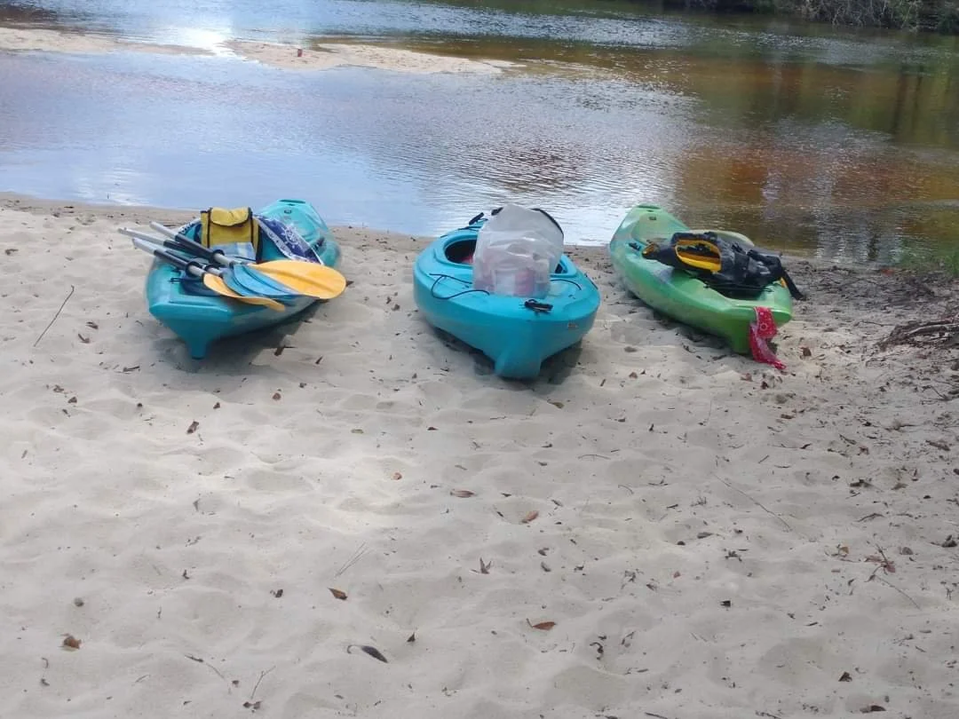 Three kayaks with paddles and gear on a sandy beach near a river.