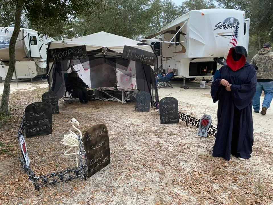 Outdoor Halloween display with gravestones, skeleton decoration, and a person in a dark robe with a red hood. There is a tent behind the display and RVs in the background, with other people nearby.