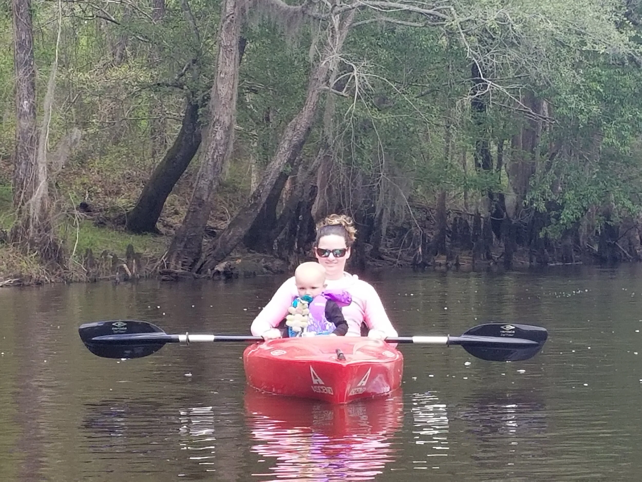 Woman in sunglasses smiling in red kayak with baby in life vest holding a pacifier on a calm river with trees in the background.