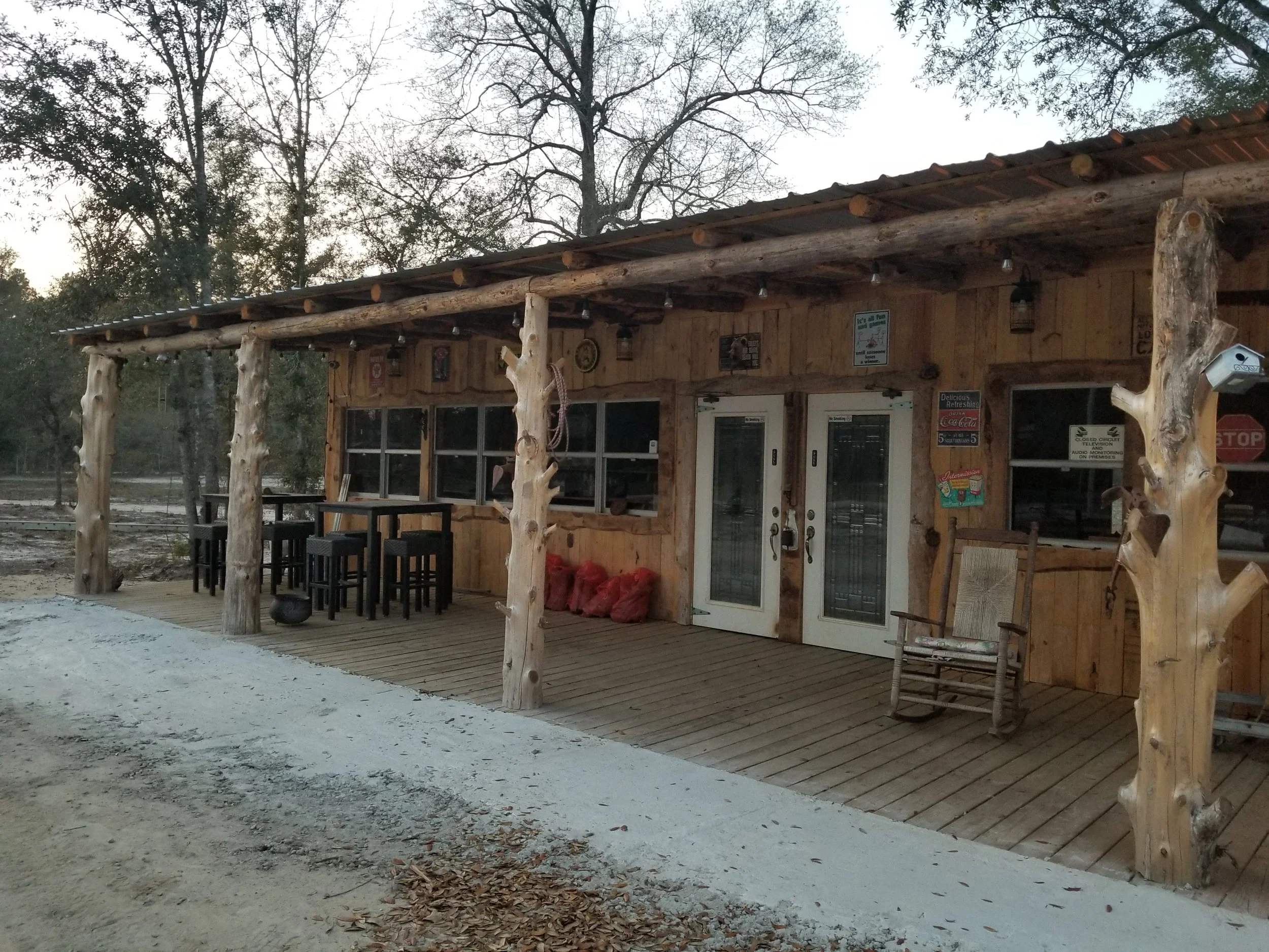 A rustic wooden building with a covered porch, outdoor tables and chairs, surrounded by trees and dirt ground.