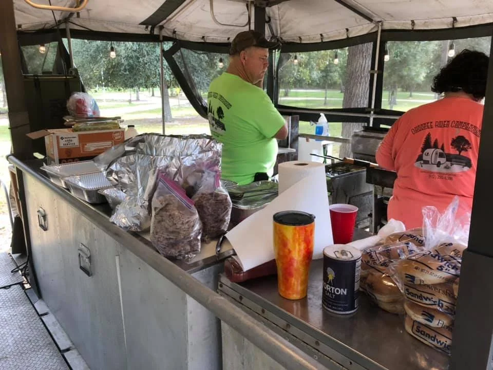 Food stall with packaging supplies, drinks, and two people working, outdoors under a canopy in a park.