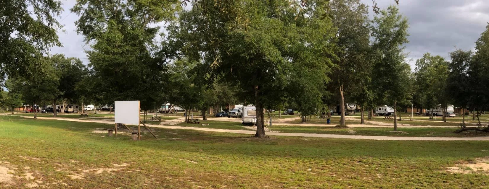 A campground with several RVs parked among tall green trees, walking paths, picnic tables, and a large blank white sign.