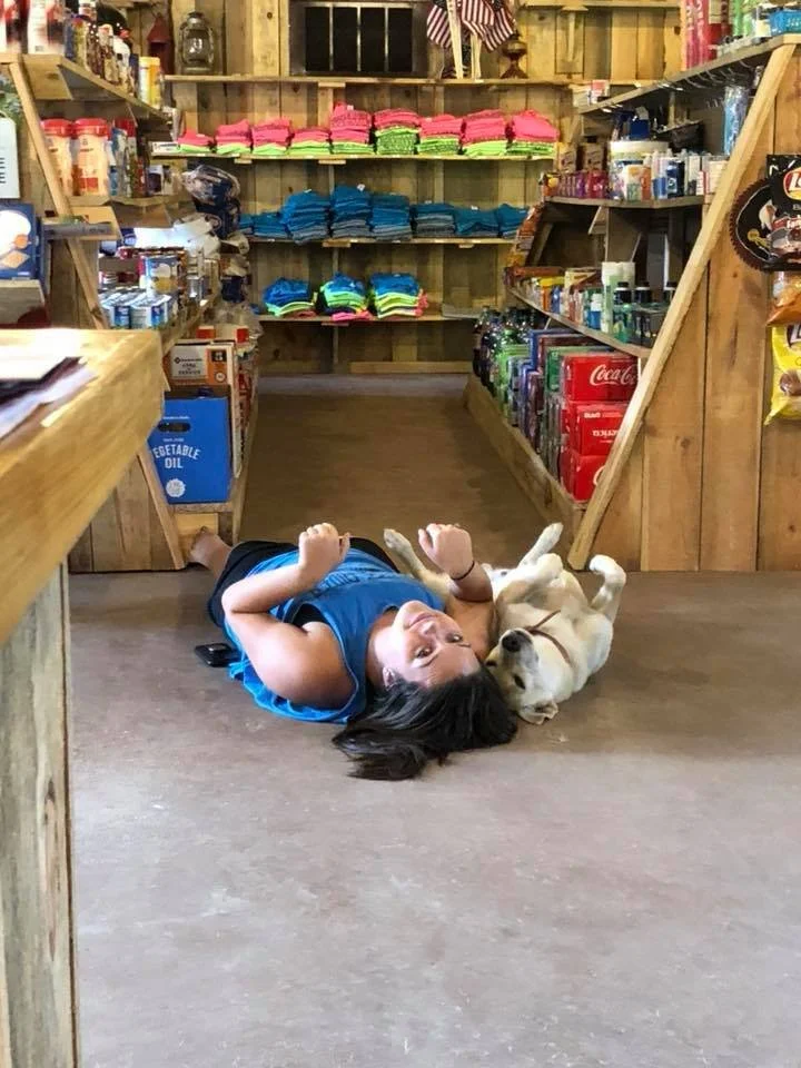 A young girl and a small dog lying on the wooden store floor underneath wooden shelves stocked with colorful folded clothes, snacks, and canned goods.