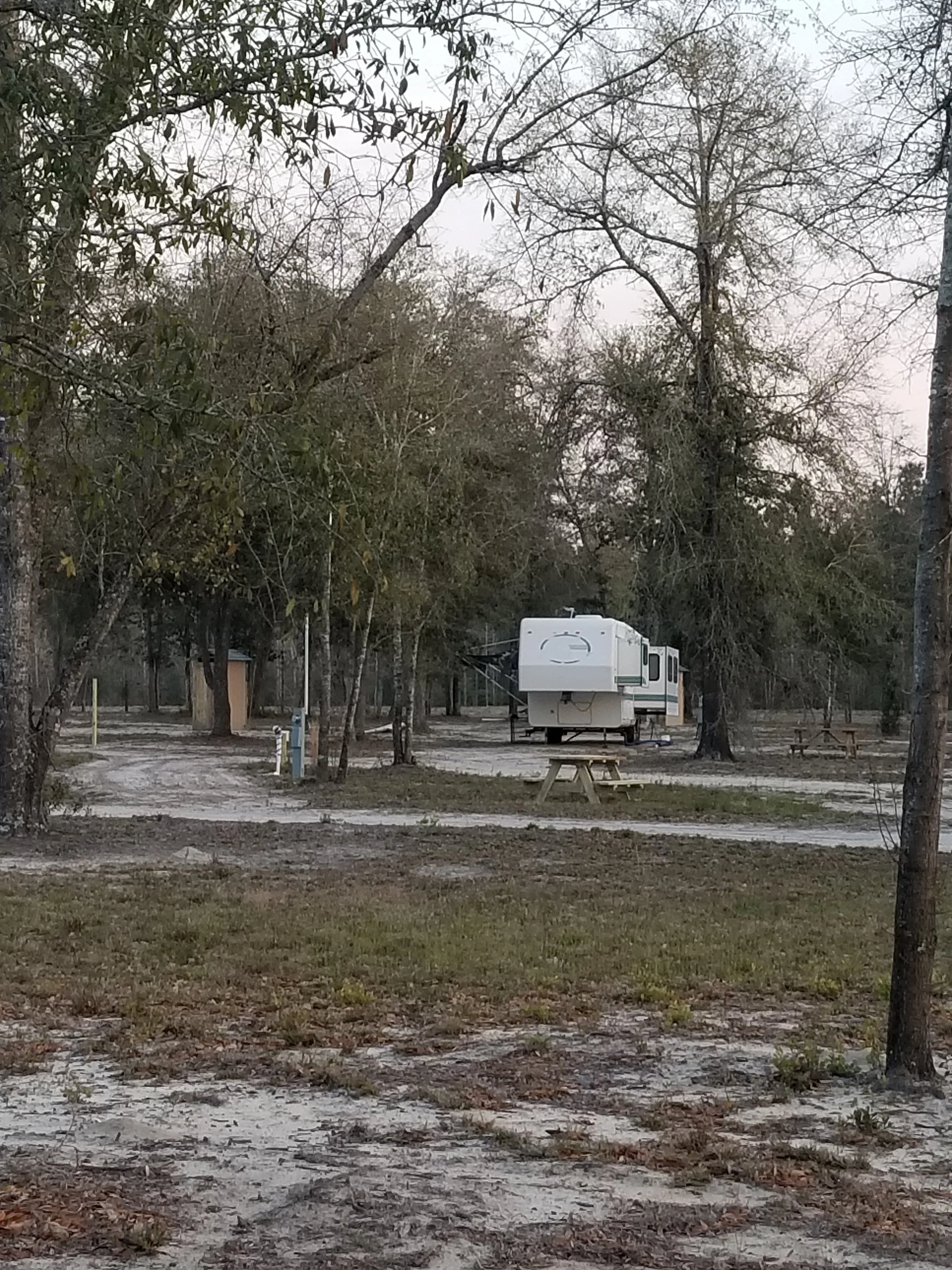 An outdoor campsite with a white travel trailer parked among trees, with picnic tables and a gravel pathway.