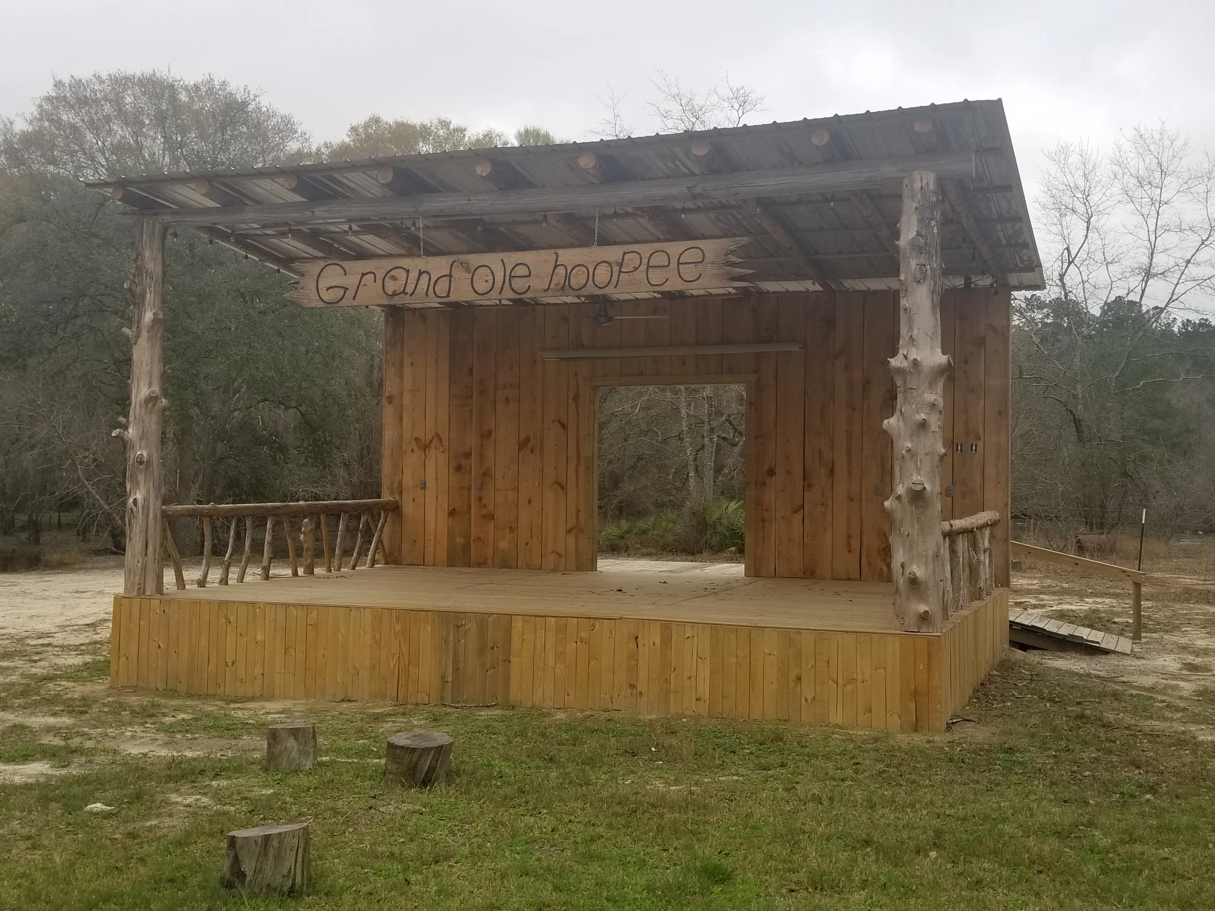 A wooden outdoor stage with a roof, surrounded by trees and grass. There is a hand-painted sign saying "Grand Ole Hoopoe" hanging from the roof.