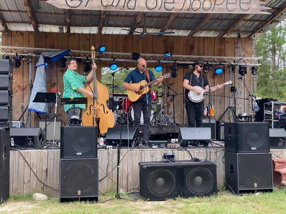 Three musicians performing on an outdoor wooden stage with a sign overhead that reads 'Hope.' The band includes a female double bassist, a male guitarist, and a female banjo player. There are musical equipment and speakers on stage.