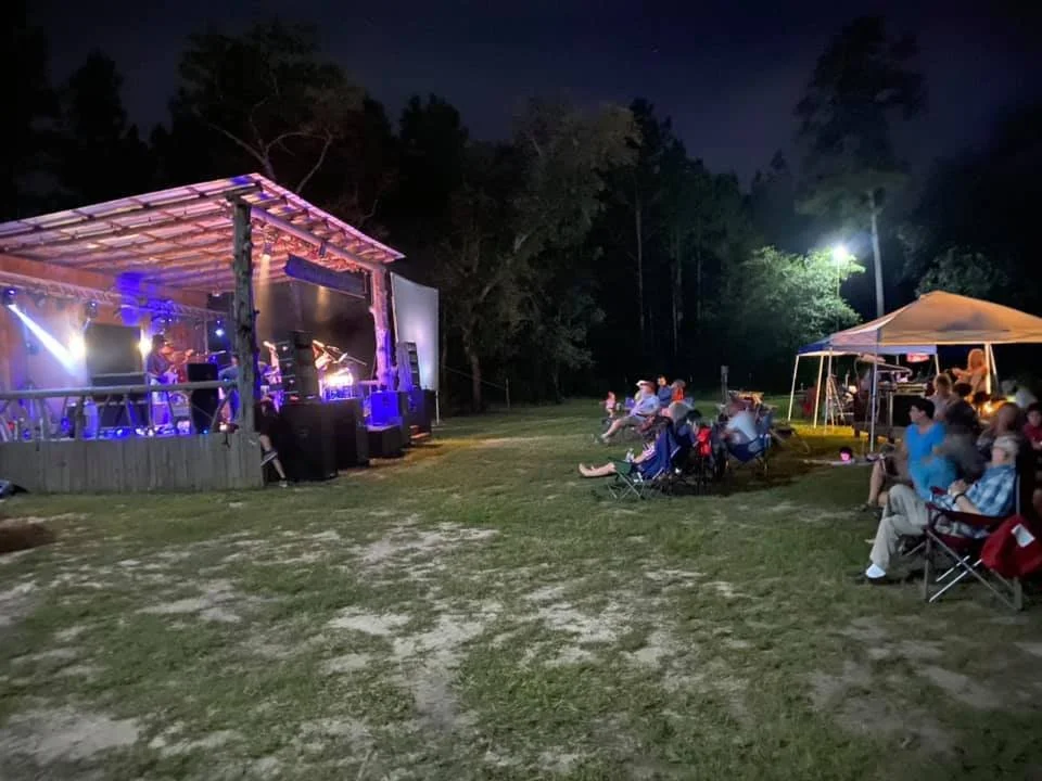 Outdoor concert at night with a stage on the left playing music and an audience seated on lawn chairs on the right under a white canopy, surrounded by trees.