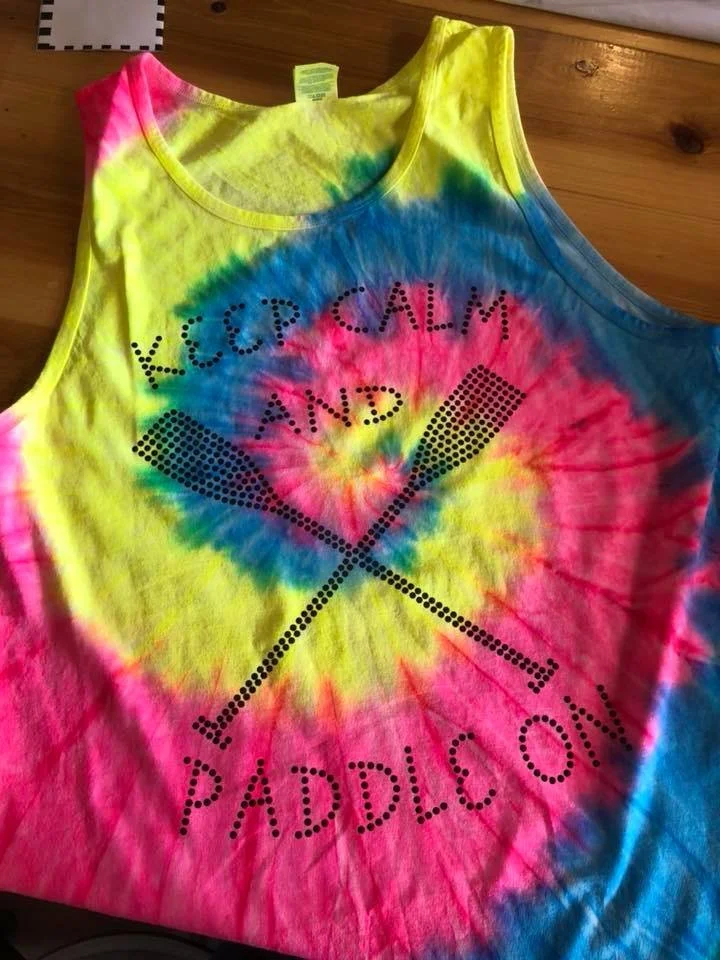 A colorful tie-dye tank top with yellow, pink, blue, and green swirls. The shirt features black dotted text that reads 'Keep Calm and Paddle On' with a crossed paddles graphic.