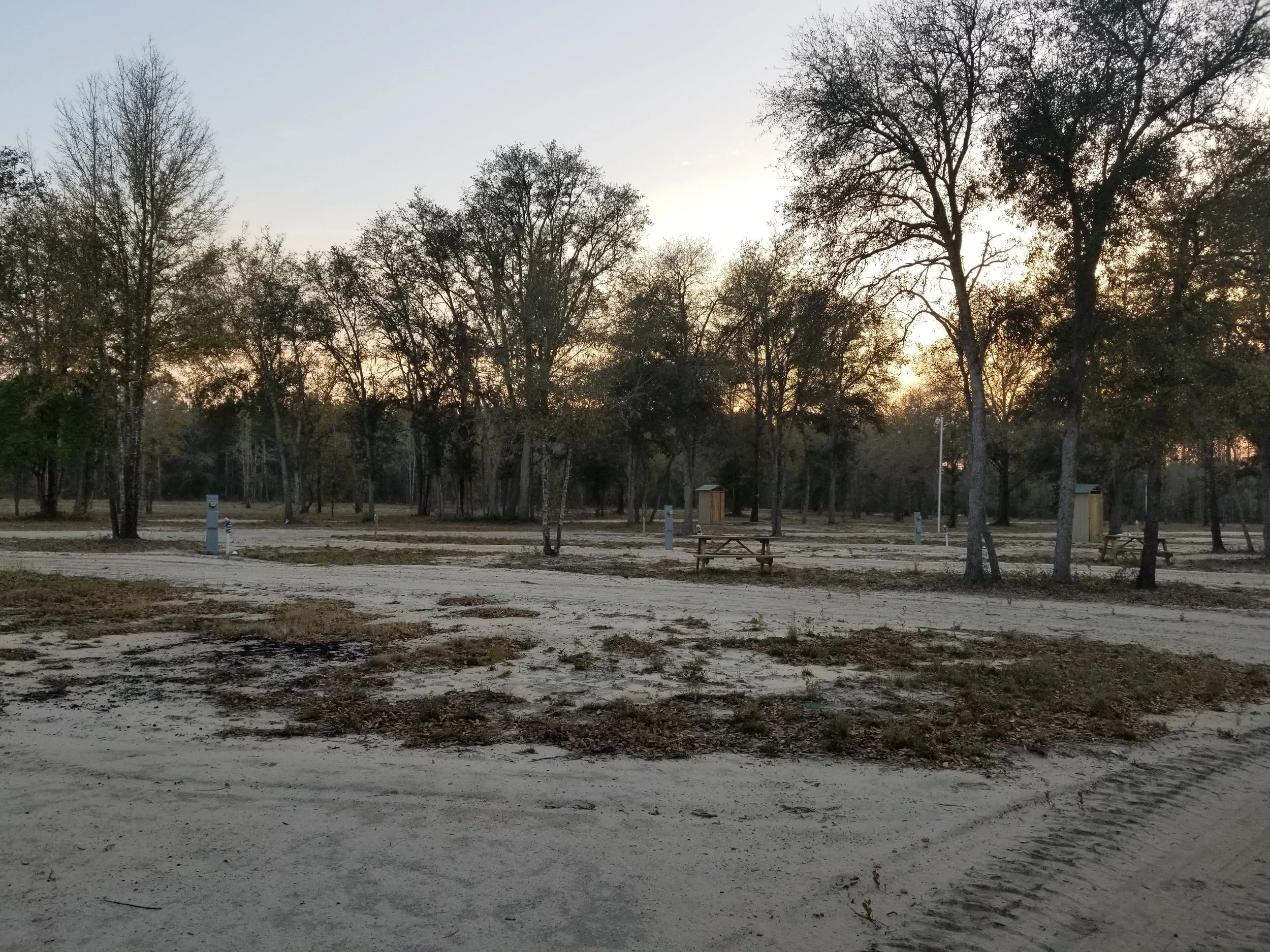 A sandy outdoor campground area with trees, picnic tables, and small structures at sunset.