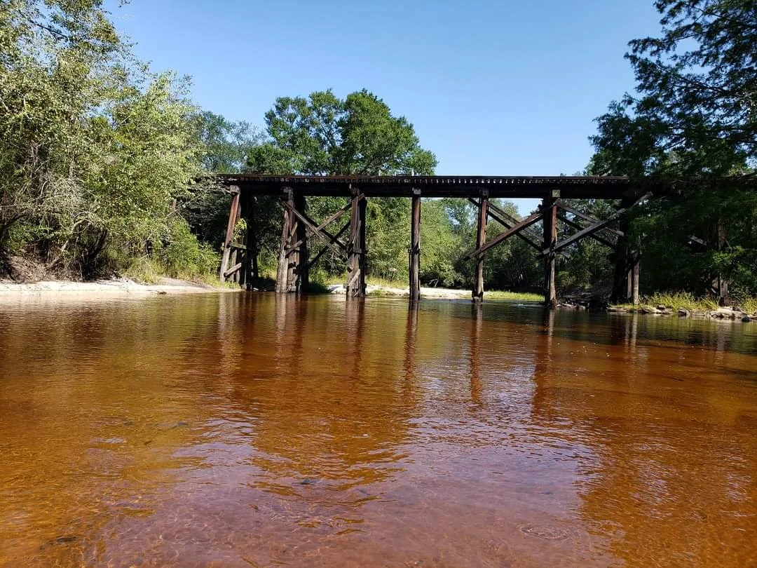 Old wooden train trestle spanning over the calm Ohoopee River with green trees and a clear blue sky.