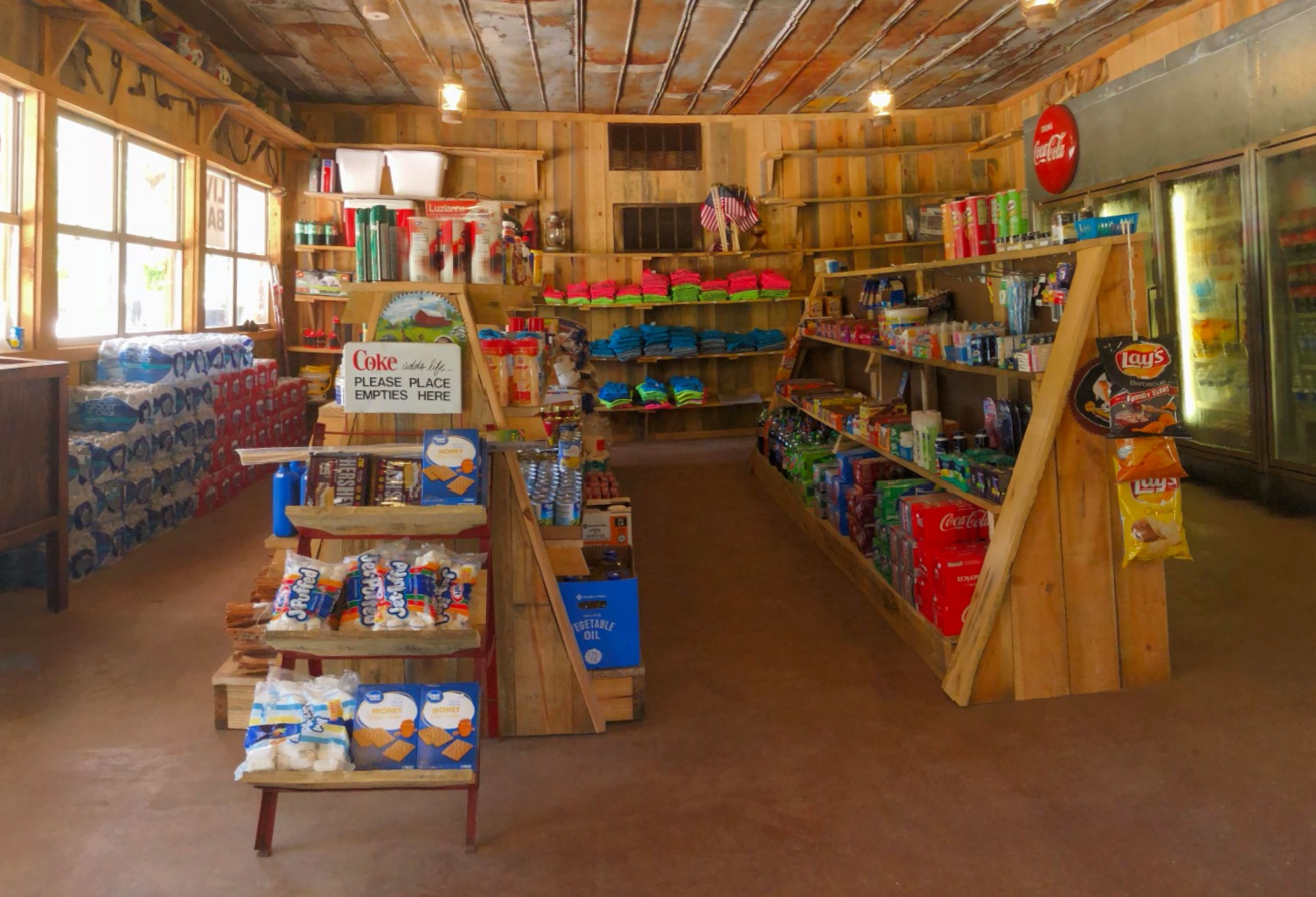 Interior of a small convenience store with wooden shelves stocked with snacks, drinks, and colorful clothing. There are windows on the left side allowing natural light in, and a Coca-Cola sign is visible on the upper right. A sign in the middle reads