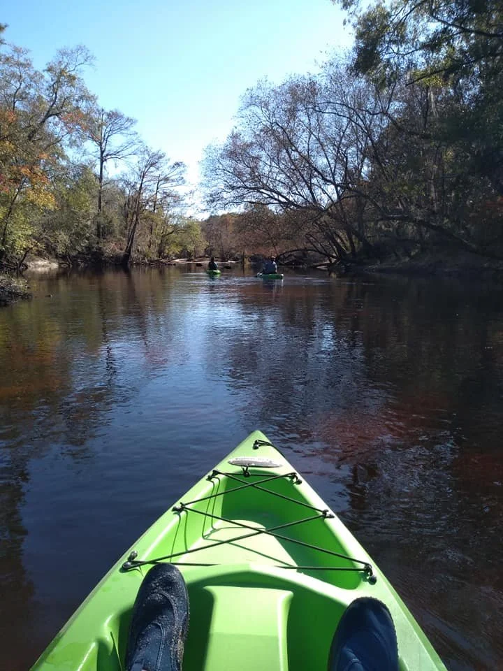 View from a kayak on a calm river surrounded by trees, with two other kayakers in the distance under a clear sky.