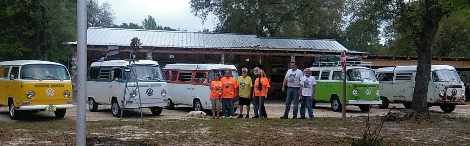 Seven people standing in front of a row of colorful vintage Volkswagen vans in an outdoor setting with trees and a rustic building in the background.