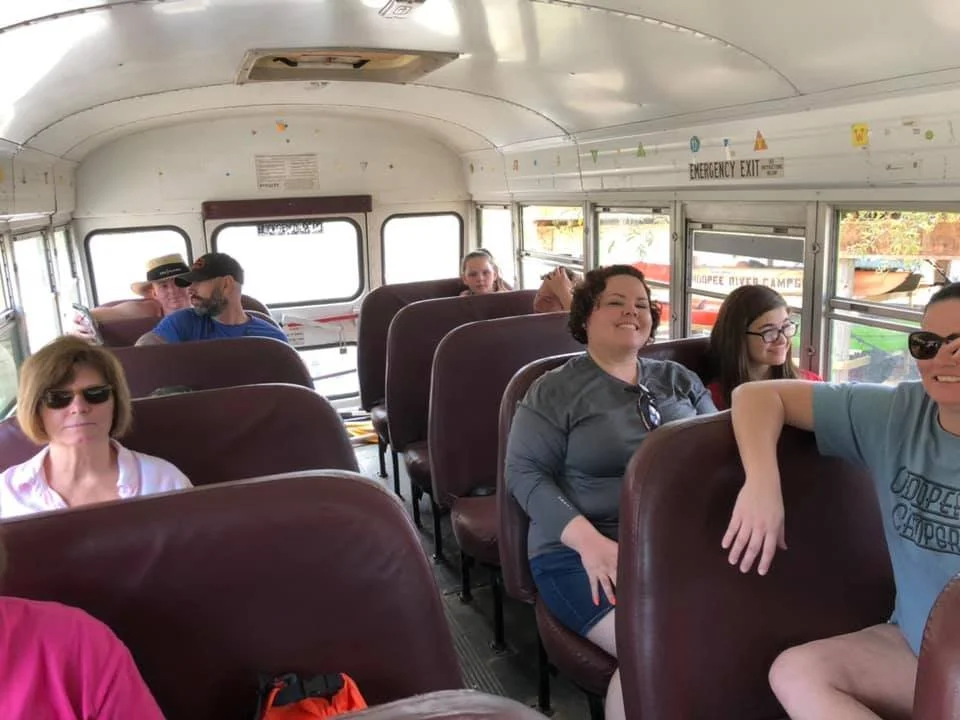 People sitting on a school bus, some wearing sunglasses, some smiling, with windows showing a sunny outdoor scene.