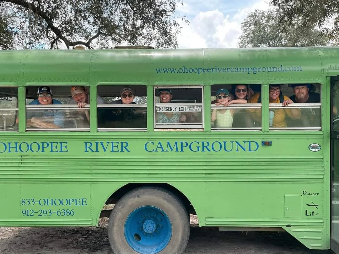 A group of people smiling and looking out from the windows of a green shuttle bus labeled 'Ohoopee River Campground'.