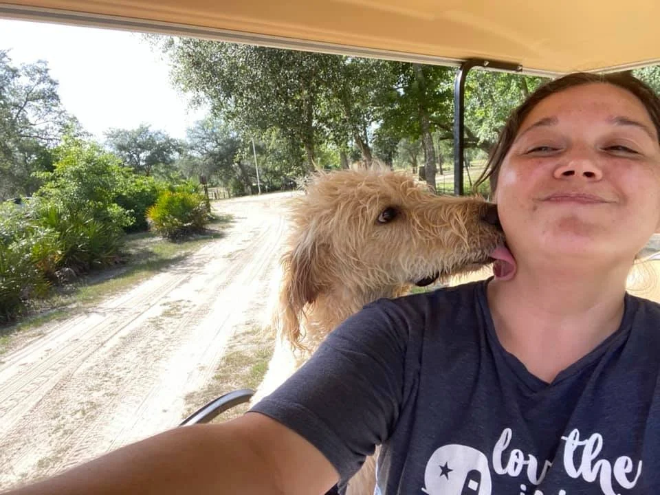 A woman taking a selfie with a light-colored dog licking her face on a sunny outdoor path with trees and bushes in the background.