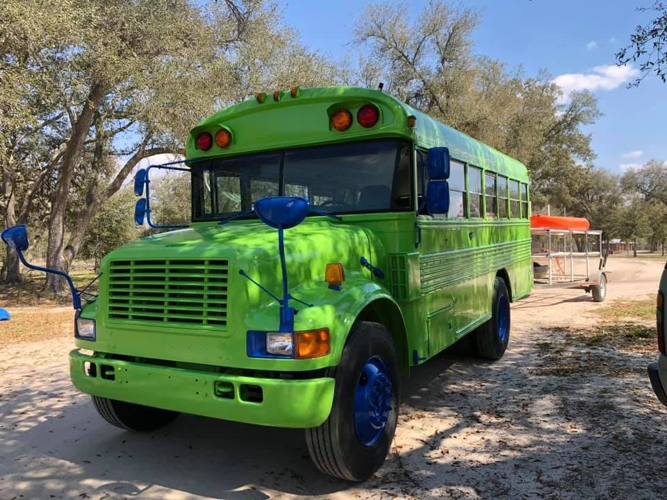 A colorful shuttle bus painted bright green with blue mirrors, wheels, and accents, parked outdoors with trees in the background.