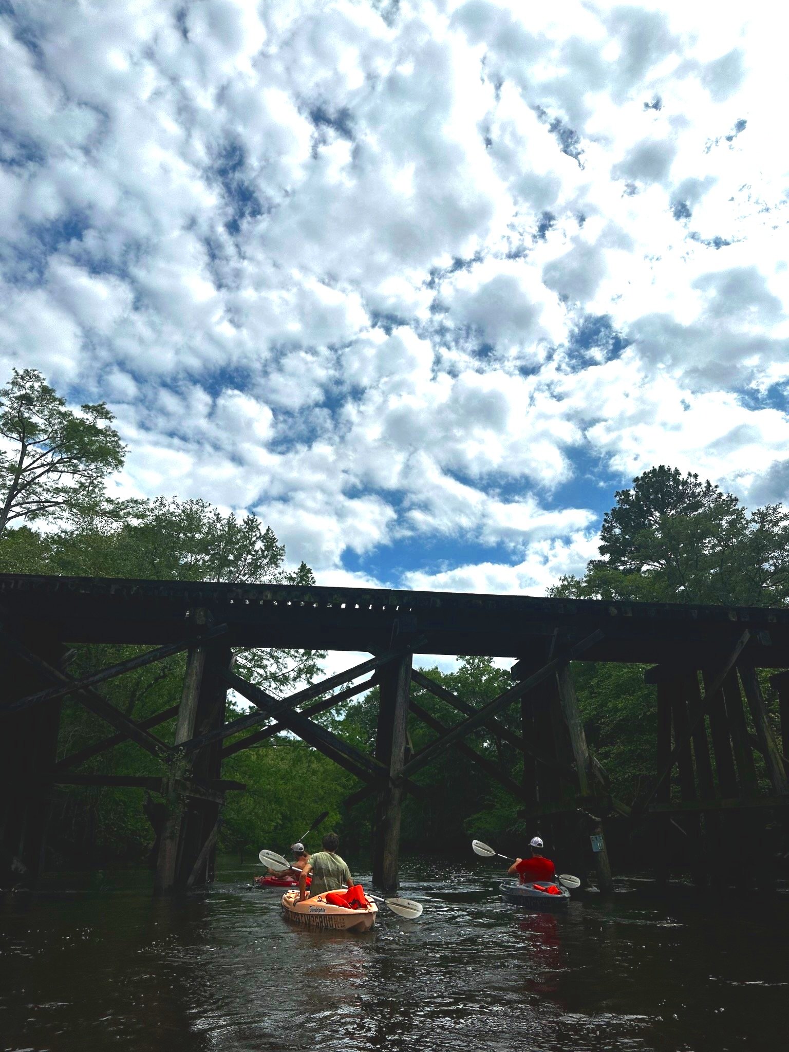 Three people kayaking under a wooden bridge on a river with a partly cloudy sky above.
