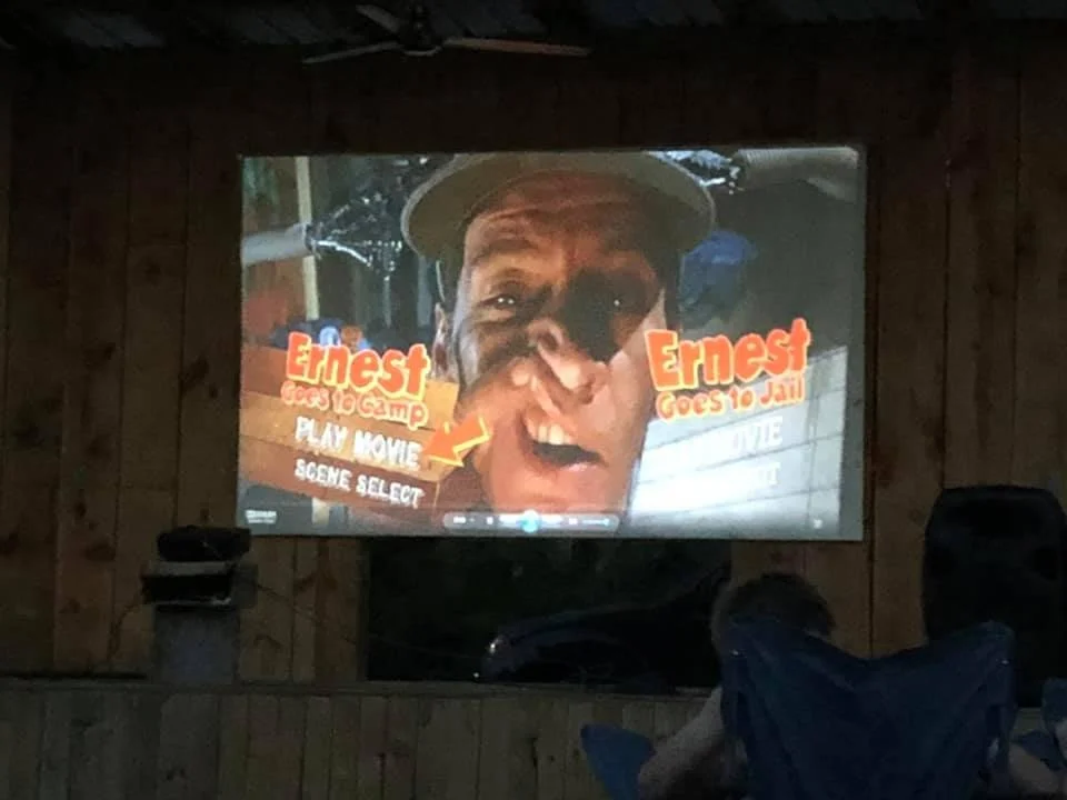 DVD menu screen for the movie "Ernest Goes to Camp" with options to play the movie or scene select. The screen features a close-up of Ernest, a man with a playful expression, wearing a cap.