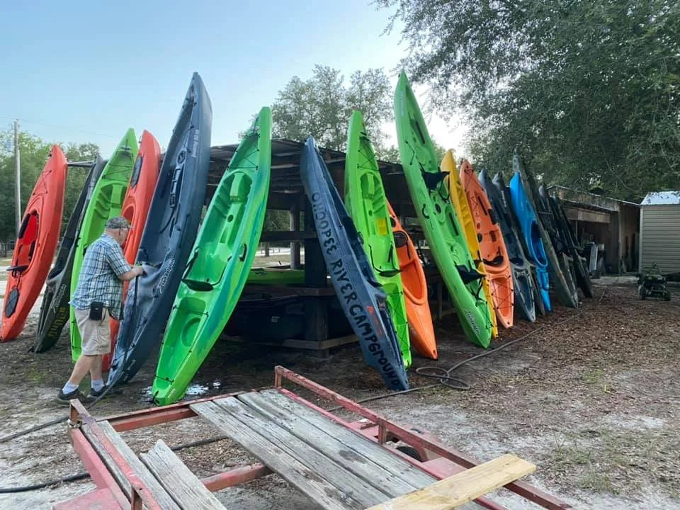 A man in a checkered shirt and shorts cleaning kayaks of various colors, including red, green, blue, and orange, which are stacked upright outside a shed.