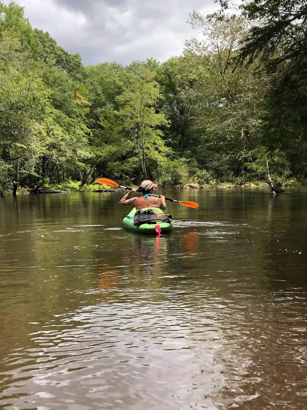 Person kayaking down a river surrounded by green trees with overcast sky.